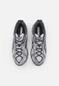 Par športni tekaški čevlji New Balance v sivi in beli barvi, izdelani iz mrežaste tkanine in z nubukastimi prekrito, prikazani od zgoraj na beli podlagi.