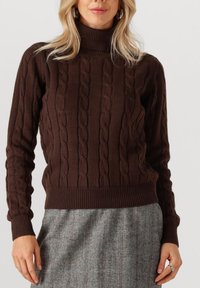 Bruine gebreide trui met een hoge hals, voorzien van een kabelpatroon en geribde mouwen en zoom. G gedragen met een grijze rok met een herringbone-patroon.