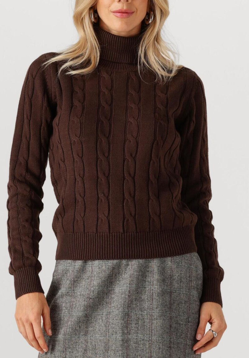 Bruine gebreide trui met een hoge hals, voorzien van een kabelpatroon en geribde mouwen en zoom. G gedragen met een grijze rok met een herringbone-patroon.