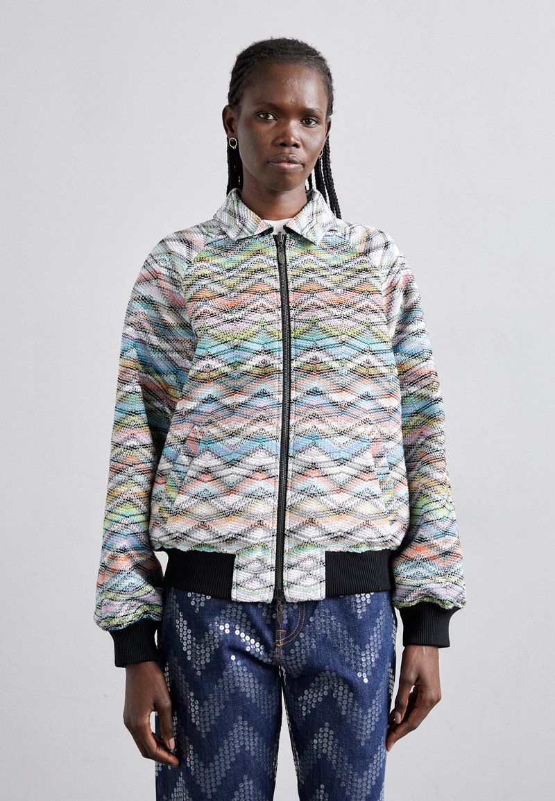 Veste bomber multicolore avec un motif en zigzag, diverses textures, poignets côtelés et une fermeture éclair à l'avant. Associée à un jean bleu marine pailleté.