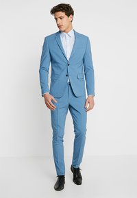 Traje azul claro hecho de tela suave. Chaqueta ajustada con dos botones, pantalones a juego y una camisa azul clara debajo. Zapatos negros.