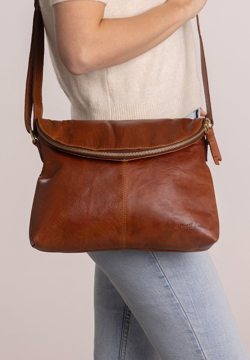 Brown leather crossbody bag with a zip closure, flat design, and a textured finish. Features an adjustable strap and a subtle logo detail.