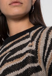 Partie inférieure du visage et cou d'une personne portant des boucles d'oreilles créoles et un pull en tricot rayé noir, beige et blanc avec un col côtelé.