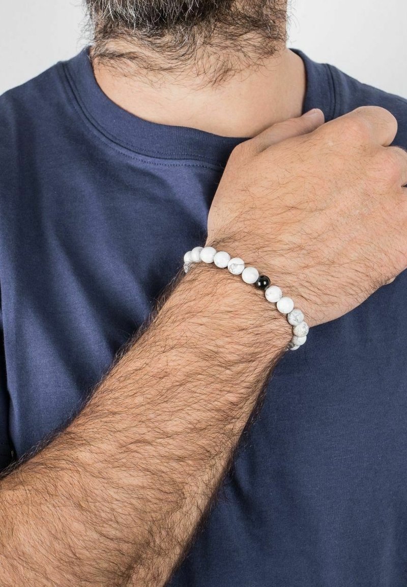Personne barbu portant une chemise bleu marine avec un bracelet de perles en marbre blanc comportant une perle noire au poignet.