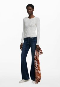 Maglietta bianca a maniche lunghe con motivo testurizzato, abbinata a jeans svasati blu scuro. Il modello tiene una sciarpa patterned arancione e marrone.