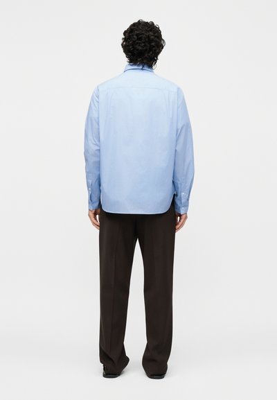 Άνδρας με σγουρά μαλλιά, που φορά ένα ανοιχτό μπλε πουκάμισο με μακριά μανίκια και σκούρα καφέ παντελόνια, στέκεται πλάτη σε ένα απλό λευκό φόντο.