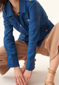 Blå linneskjorta med två framfickor, rundad krage och träknappar. Modellen kombinerade den med bruna vida byxor och sandaler med remmar.