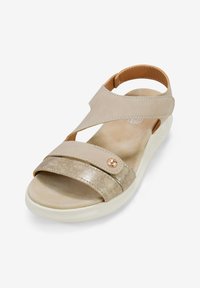 Sandal beige con cinturino metallico dorato, design incrociato, cinturino alla caviglia regolabile, soletta morbida e dettaglio decorativo con bottone metallico.