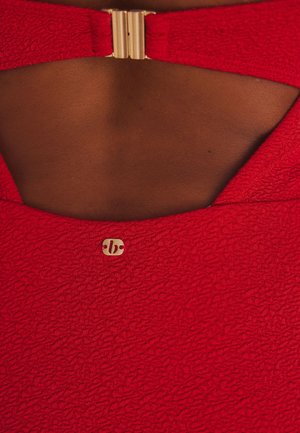 Parte trasera de piel oscura visible a través de un corte en tela roja texturizada con pequeño broche dorado y detalle de logo dorado debajo del corte.