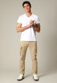 Witte V-hals T-shirt gecombineerd met beige cargobroek met ritszakken. Schoenen zijn witte sneakers met zwarte accenten.