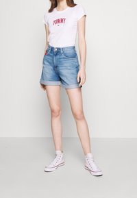 White t-shirt with red "TOMMY" print, paired with light blue denim shorts featuring rolled cuffs, worn with white high-top sneakers.