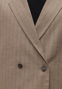 Blazer marron clair à rayures fines avec de larges revers et deux boutons foncés sur un vêtement intérieur noir.