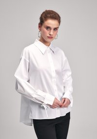 Camicia bianca a maniche lunghe con colletto classico, dotata di polsini con cravatta regolabili e orlo posteriore più lungo. Indossata con pantaloni neri.