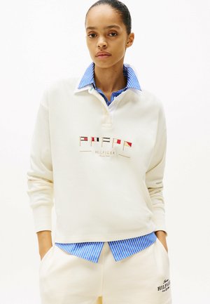 Mujer con sudadera color crema con banderas náuticas y el logo "Hilfiger", sobre una camisa de cuello a rayas azules, con las manos en los bolsillos.