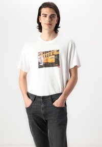 Tricou alb din bumbac, cu un grafic colorat de graffiti pe un perete, asortat cu blugi gri închis. Croială simplă cu guler rotund și mânecă scurtă.
