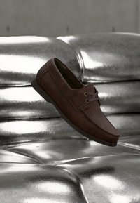 Mocasín de cuero marrón con cordones oscuros descansando sobre un tapizado plateado brillante y capitoné.