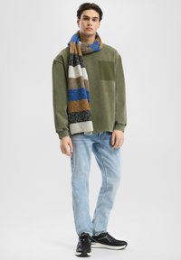 Olivovo zelený sveter s vreckom na hrudi, svetlomodré džínsy a viacfarebný pruhovaný šál. Čierne športové topánky dopĺňajú celý outfit.