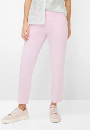 Kvinde iført lyserøde, tætsiddende bukser, beige sneakers og en hvid, tekstureret bluse mod en ensfarvet hvid baggrund.