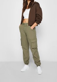 Cargo pants in olive green with multiple pockets, elastic cuffs, and a zip fly, paired with a cropped brown hoodie and white sneakers.