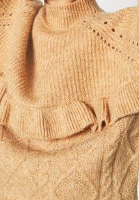 Gros plan d’un pull beige doux en tricot avec un col montant et un détail froncé au niveau de la poitrine et de la manche.