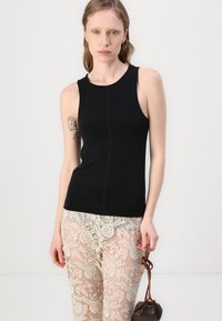 Haut sans manches noir avec un col rond, présentant une coupe ajustée. Associé à un pantalon transparent avec un motif en dentelle et un petit sac marron.