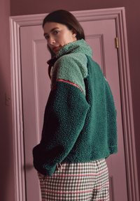 Merdkangast pullover türkiissinistes ja rohelises toonides, kõrge kaeluse, tekstuurse pinnaga ning roosade aksentidega, kombineeritud ruuduliste pükstega, millel on roheline ja valge värv.