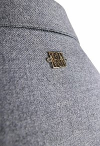 Szary, teksturowany materiał z metalowym logotypem w kolorze złotym, zdobionym misternym wzorem. Lekko podniesione przeszycia wzdłuż górnej krawędzi.