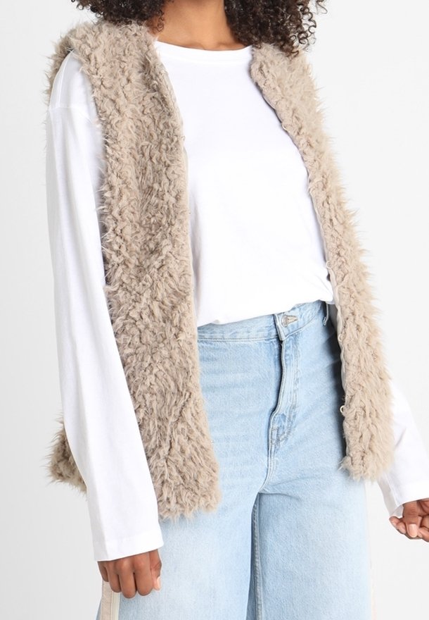 Faux fur-väst i beige med en texturerad, fransig yta. Öppen frontdesign, kombinerad med en vit långärmad skjorta och ljusblå jeans.