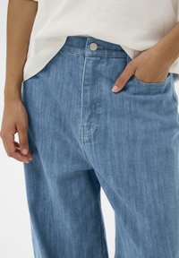 Svetlo modri kavbojki s sproščenim krojem, ki imajo sprednji žep in srebrno gumbe podrobnost na pasu.