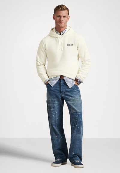 Muž v krémovej mikine s kapucňou cez modrú pruhovanú košeľu, v roztrhaných džínsach s nášivkami a v námornícky modrých teniskách bez šnúrok, stojaci pred jednoduchým bielym pozadím.