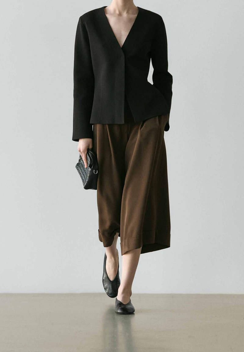 Personne portant une veste noire à col en V, un pantalon marron large et court, des ballerines noires, et tenant une petite pochette noire.