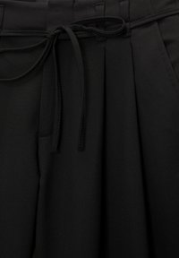 Pantalon en tissu noir avec un cordon noué à la taille et des passants de ceinture ainsi que des plis visibles.