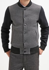 Grijze wolmix baseballjack met zwarte mouwen, geribbelde manchetten en een knopensluiting aan de voorkant met twee zijzakken.