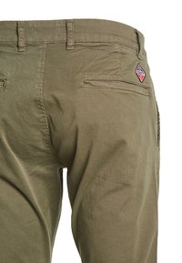Pantalones color caqui con una textura suave, que cuentan con bolsillos traseros y un parche con un logo en forma de diamante. El diseño es funcional y sencillo.