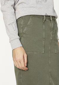 Jupe en denim vert olive avec deux poches frontales, présentant des coutures visibles. Associée à une chemise à manches longues grise ornée de dessins en ligne noirs.