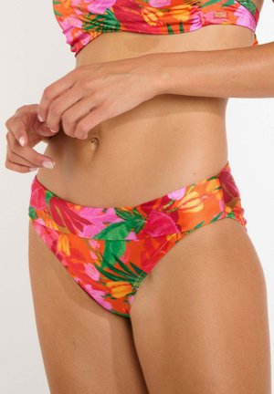 Femme portant un bas de bikini floral éclatant et un haut assorti, mains près de la taille, montrant la taille et le teint de la peau.