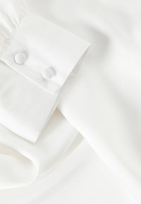 Bela bluza z gladko teksturo, z manšetom, ki ima dve gumbi iz istega blaga, in nežno padajočim tkanjem pod manšetom.