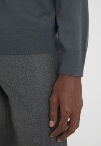 Maglione grigio a maglia con orlo a costine, abbinato a pantaloni grigi scuri con texture. L'immagine mostra un primo piano della manica e della mano.