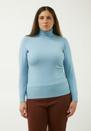 Un pull col roulé bleu clair en tissu doux, avec des poignets et un ourlet côtelés, porté avec un pantalon texturé marron.