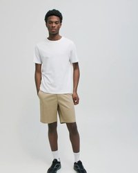 Noor mees seisab käed taskus, kandes valget t-särki, beeže lühikuid pükse, valgeid sokke ja musti tossusid ühevärvilise tausta ees.