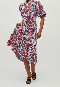 Robe fleurie à manches bouffantes avec motifs rouges, blancs et bleus. Longueur sous le genou, matériau léger, associée à des sandales noires.