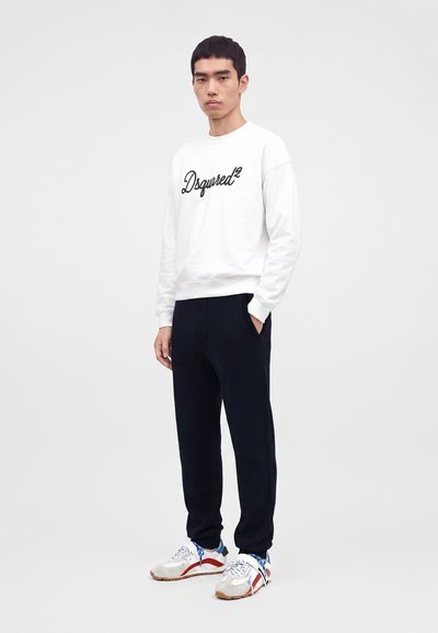 Jeune homme debout portant un sweat-shirt blanc avec un texte noir, un pantalon noir et des baskets blanches avec des accents rouges et bleus.
