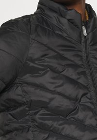 Giacca a vento nera con colletto alto, chiusura con zip e dettagli a motivo chevron sul tessuto. Texture liscia e lucida.