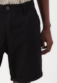 Svarta bomullsshorts med knappstängning, sidofickor, uppvikta fållar och en mjuk yta. Provanvända av hand som håller i shortsen.