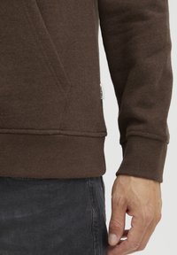 Brown sweatshirt made of soft cotton blend. Features ribbed cuffs and hem, a front pocket, and a small brand tag on the side.