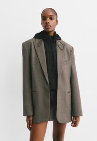 Blazer marron ajusté avec une veste intérieure à capuche noire zippée, présentant un bouton unique, des poches plaquées et une texture lisse.