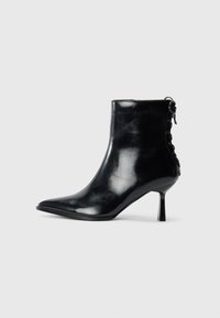 Botte de cheville en cuir noir brillant avec un embout pointu, un talon aiguille et un détail lacé à l'arrière, sur fond blanc.