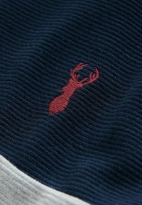 Navy zilas ribots audums ar mazu sarkanu izšūtu brieža logotipu. Mīksta tekstūra ar kontrastējošu gaiši pelēku apakšējo malu.