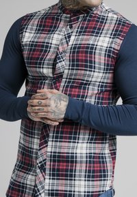 Plaid shirt with a navy long-sleeve base, featuring red, white, and blue squares, button-up design, and embroidered logo on the chest.