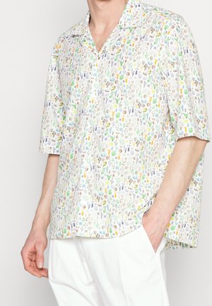 Uomo con una camicia bianca a maniche corte con piccoli motivi colorati di cactus e piante, abbinata a pantaloni bianchi.